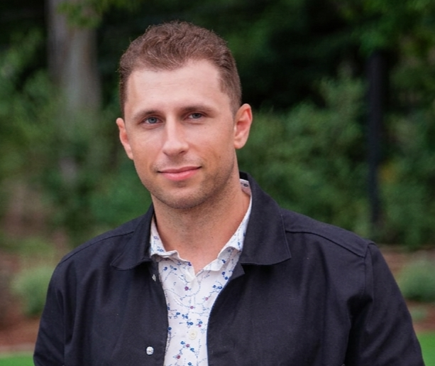 Man wearing a dark jacket over a patterned shirt with a blurred green background